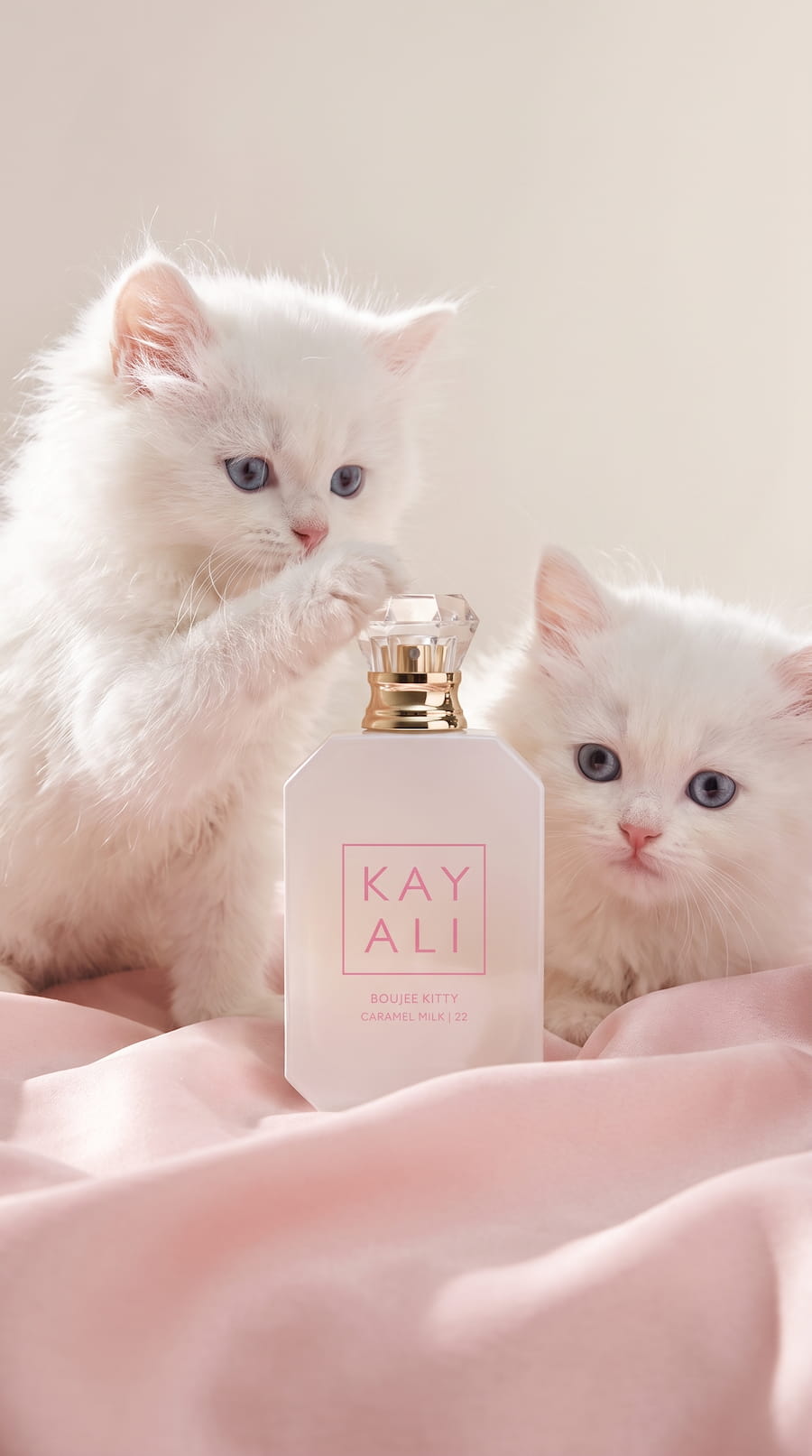 Two fluffy white kittens beside a pale pink KAYALI Boujee Kitty Caramel Milk perfume bottle on soft pink fabric.