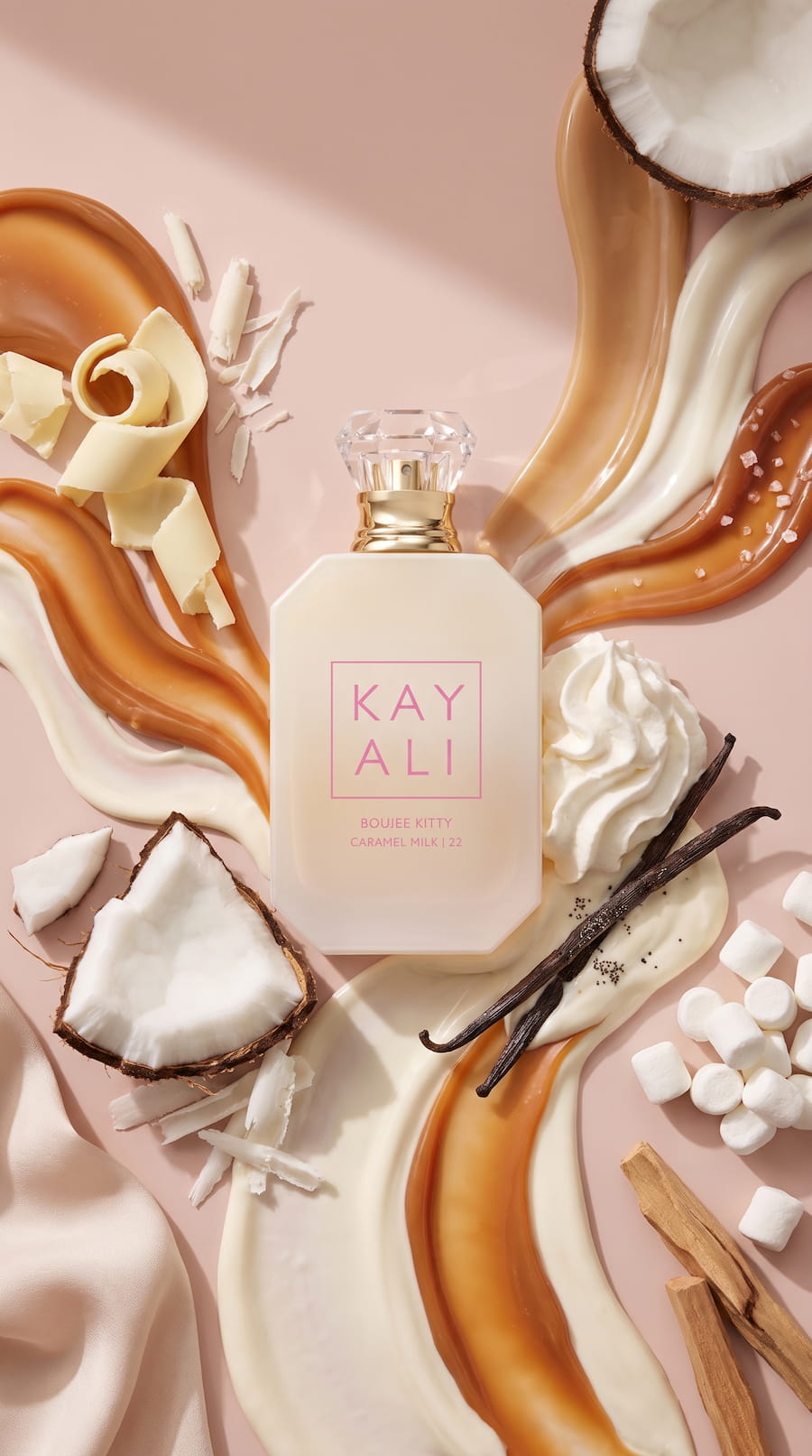 KAYALI Boujee Kitty Caramel Milk perfume bottle surrounded by caramel, cream, coconut, vanilla pods, and marshmallows on a blush pink background.
