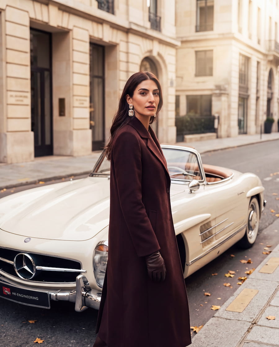 Fashion editorial image featuring a woman in a burgundy coat and statement earrings beside a vintage-style cream convertible in an elegant city setting.